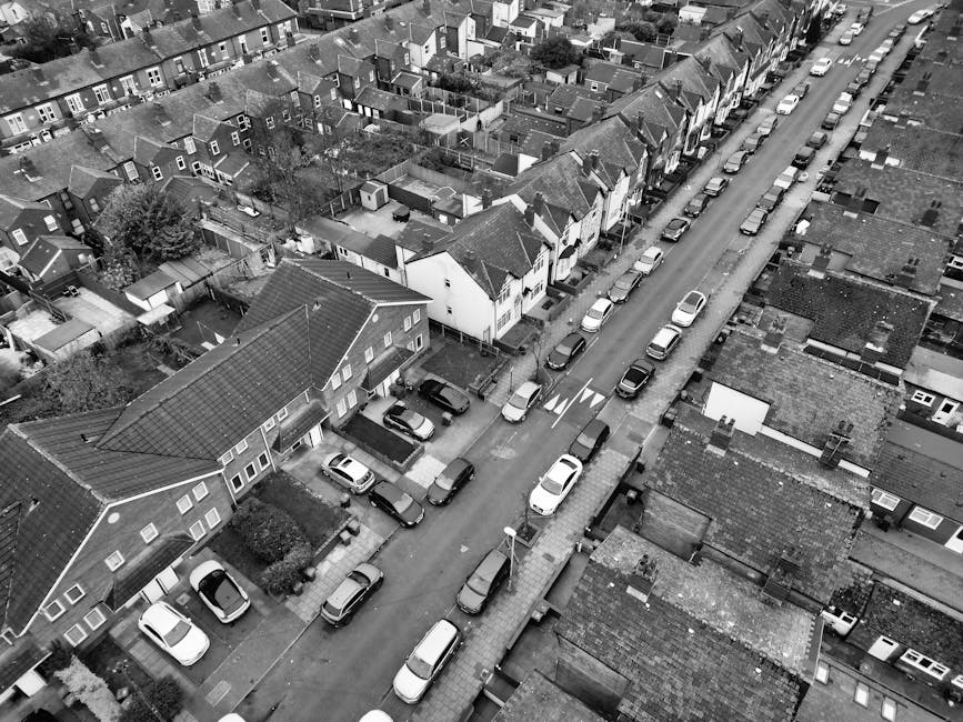 A black-and-white aerial photograph showing a residential neighborhood with narrow streets lined by closely spaced terraced houses and detached homes. On the streets, multiple parked cars are visible,