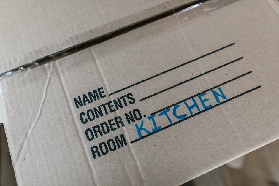 Close-up view of a cardboard moving box labeled with black printed text on a white background, featuring fields for name, contents, order number, and room, with the word 'KITCHEN' handwritten in blue