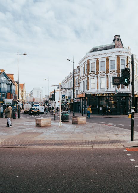 A busy street scene on West End Lane in Camden featuring a corner building with white and brown brick exterior and decorative architectural details, situated at the intersection. The foreground shows 