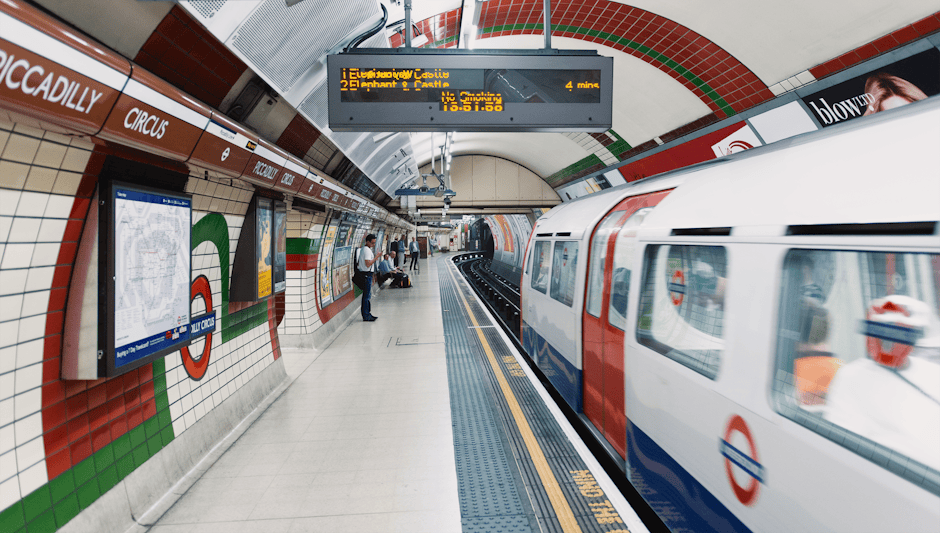 Inside a London Underground station with tiled walls decorated in red, white, green, and black patterns, a packed platform features passengers waiting for a train, with some standing and others sittin