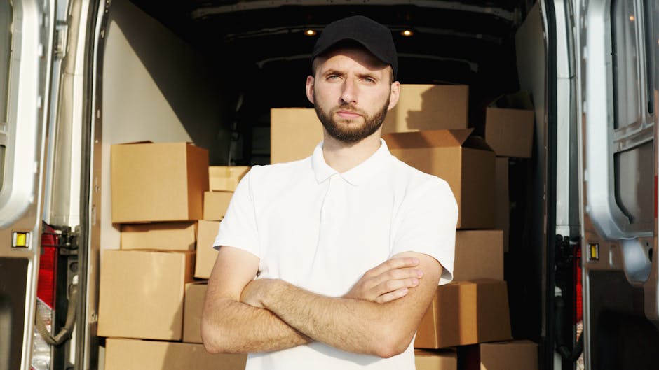 A young man with a beard, wearing a black cap and a white polo shirt, stands inside the open rear of a large moving van. The van is filled with numerous cardboard boxes of various sizes, many sealed w